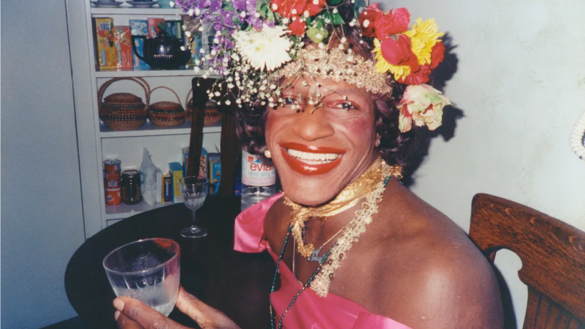Portret cu Marsha P. Johnson, activistă afro-americană transgen. Marsha poartă niște flori colorate, bijuterii și zâmbește larg.